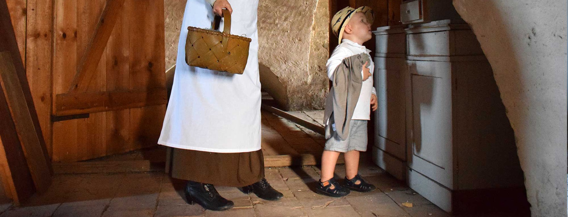 Ett litet barn iklädd stråhatt blir visad ett skåp av en traditionellt klädd person som håller en korg.